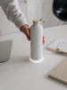 EQUA Smart Water Bottle Snow White made from Stainless Steel in White Colour and Gold Lid and Ring detail in the center of the image with hand reaching out to take it to make a sip.
